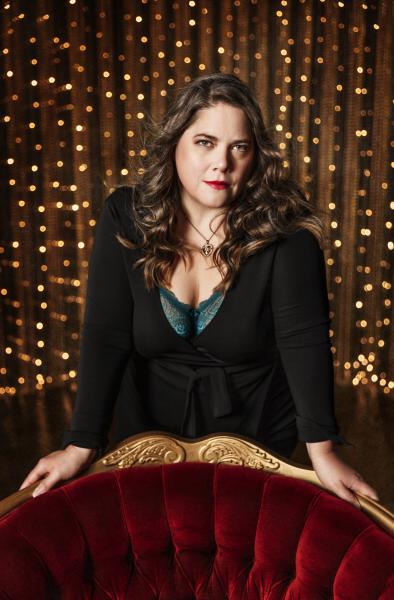 A woman with long dark hair. She is leaning on an antique chair and staring into the camera.