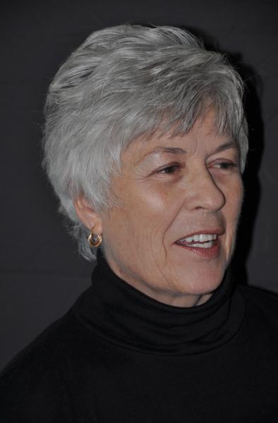 A woman with short grey hair, wearing a black turtleneck and small gold hoop earrings. She is looking off to the side and smiling slightly.