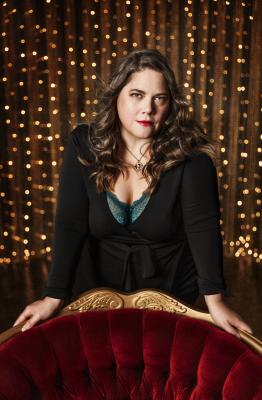 A woman with long dark hair. She is leaning on an antique chair and staring into the camera.