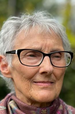 A photo of an older woman with short grey hair. She is wearing dark framed glasses and has a pink scarf around her neck. She is slightly smiling.