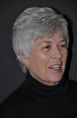 A woman with short grey hair, wearing a black turtleneck and small gold hoop earrings. She is looking off to the side and smiling slightly.